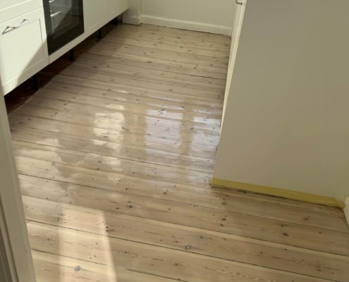 Køkken med nylakeret lyst trægulv og moderne hvide skabe.